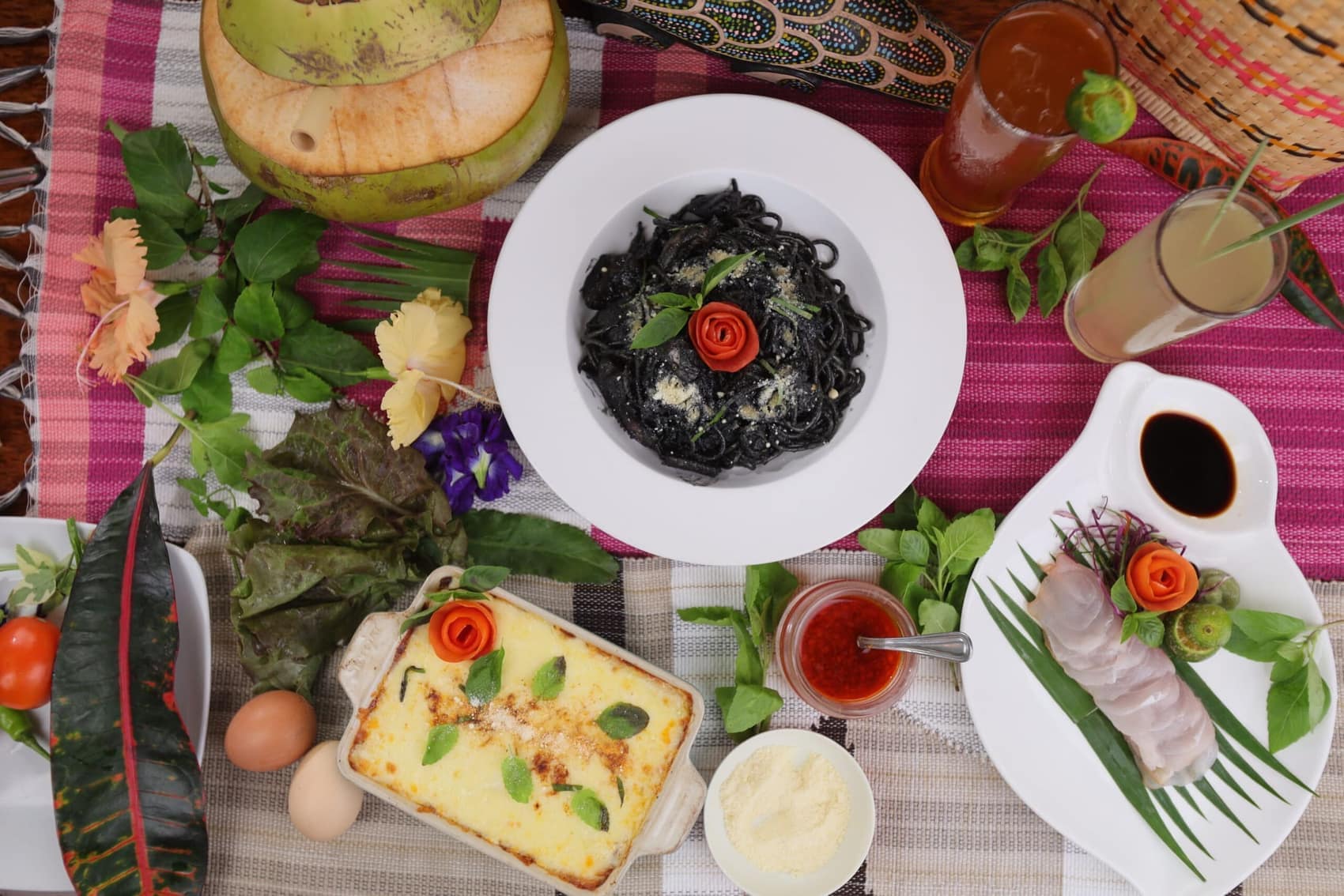 A full spread laid out on woven Palawan textiles: squid-ink pasta, lasagne, sashimi and buko juice