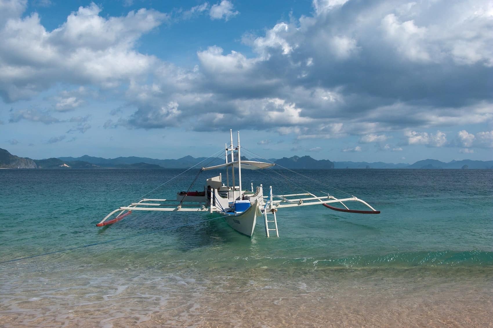 A traditional double-outrigger bangka riding at anchor in glass-clear shallows, Bacuit Bay